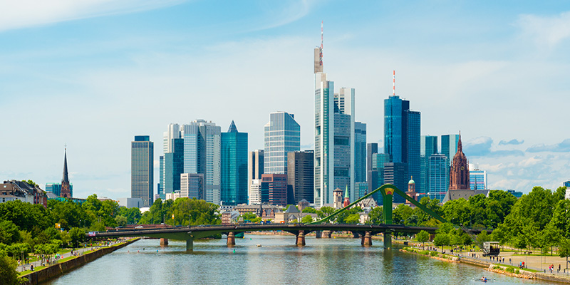 Blick auf den Main in Frankfurt mit einer Brücke und der markanten Skyline aus zahlreichen Hochhäusern im Hintergrund unter blauem Himmel